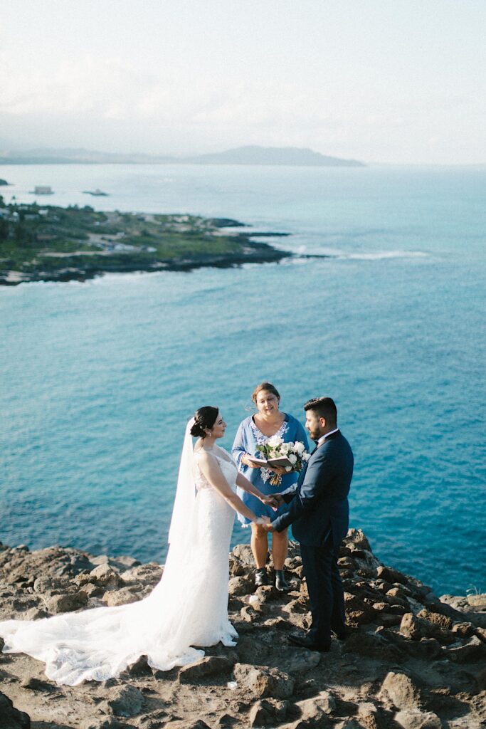 A bride and groom hold hands during their elopement ceremony on a cliff overlooking the ocean and the Hawaiian islands