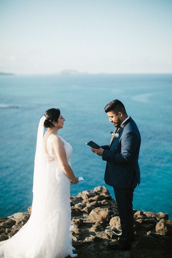 A groom reads his vows to his bride while they stand together on a cliff looking out over the Hawaiian islands and the ocean