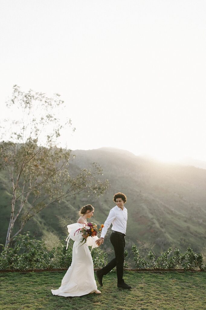 A bride and groom walk hand in hand after their elopement with mountains in the background.