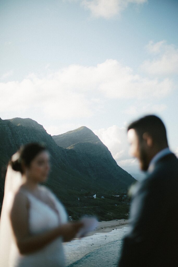 A bride and groom are out of focus in the photo reading their vows to one another, in the background are the mountains of Hawaii in focus.