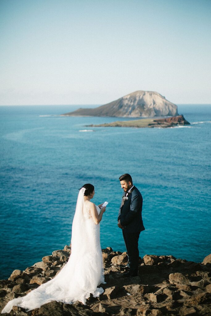 A bride reads her vows to the groom while they stand on a cliff looking out over the ocean and an island in the Hawaiian islands.