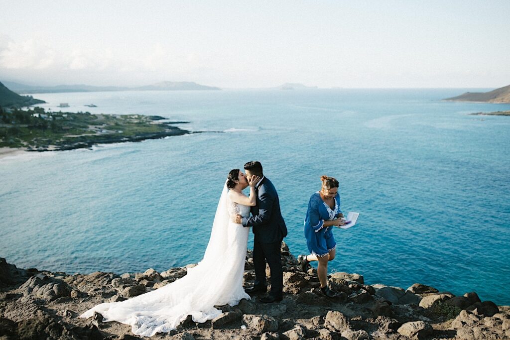 A bride and groom kiss as their officiant exits their elopement ceremony on a cliff looking over Makapuu Beach