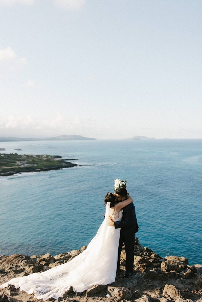 A bride and groom kiss atop a cliff looking out over the Hawaiian islands and the ocean after their elopement ceremony.