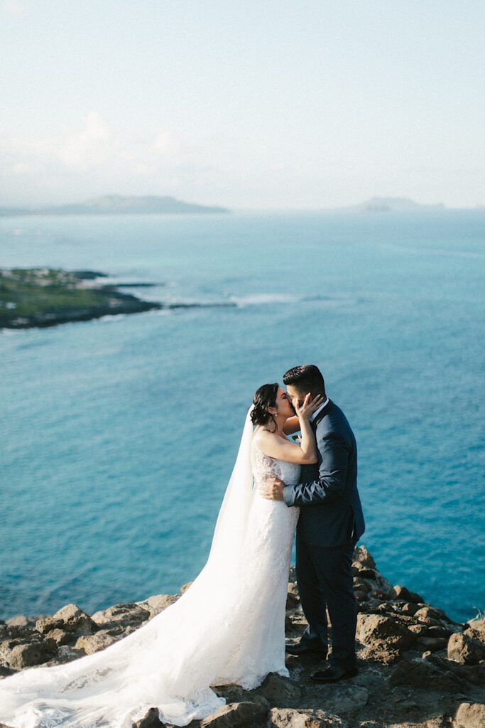 A bride and groom kiss atop a cliff looking out over the Hawaiian islands and the ocean after their elopement ceremony.