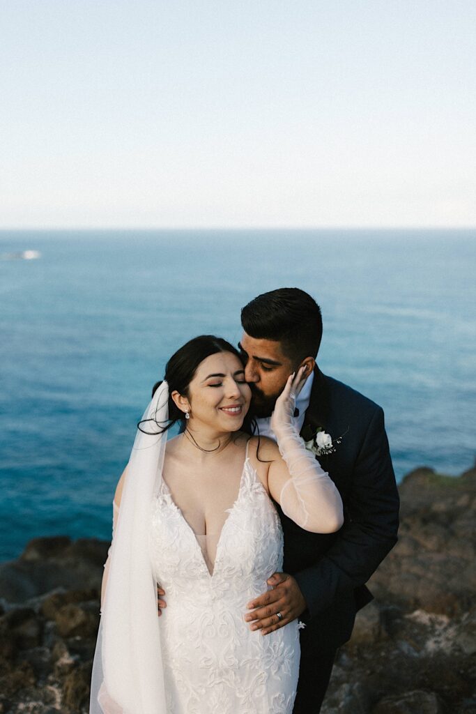 A bride and groom stand together atop a cliff overlooking the ocean of Hawaii, the groom is behind the bride kissing her on the cheek