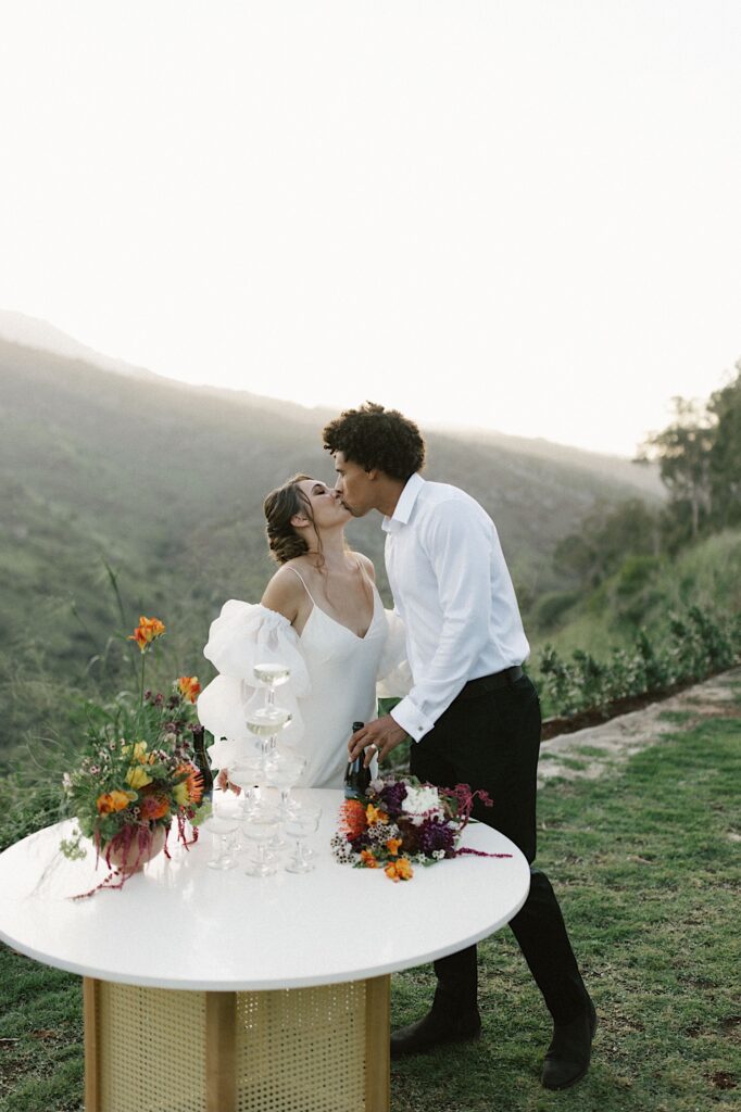 A bride and groom kiss in front of mountains, in front of them is a table with a tower of glasses on it.