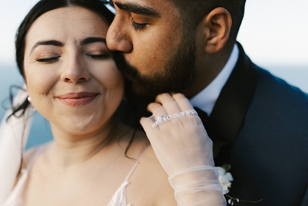 Closeup photo of a groom kissing his bride on the cheek from behind as the touches his beard.
