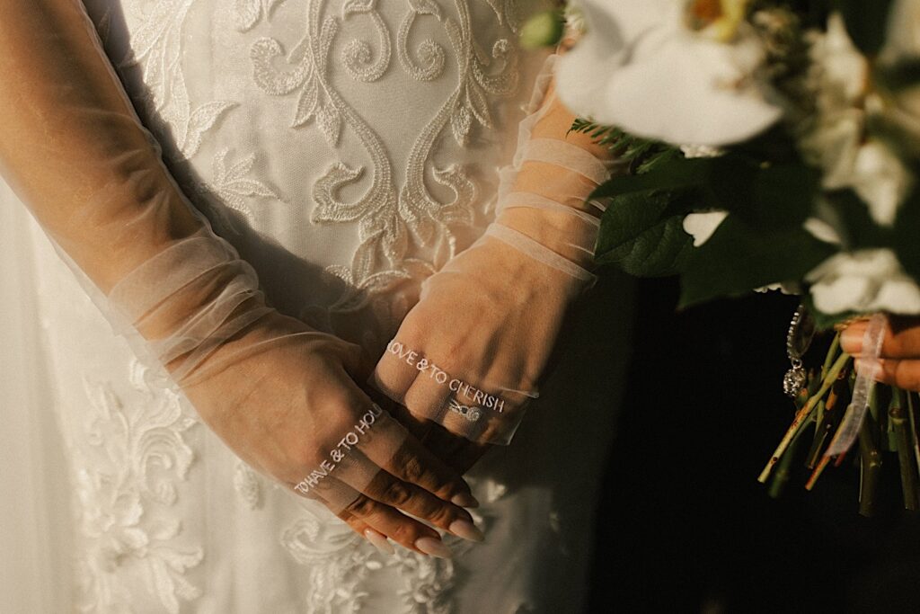 Closeup photo of a brides hands with sheer sleaves, on the sleaves are the words "to have & to hold" "to love & to cherish"