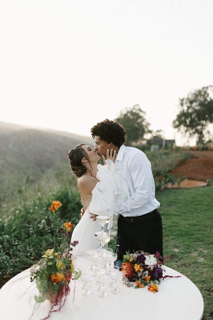 A bride and groom kiss in front of mountains, in front of them is a table with a tower of glasses on it.