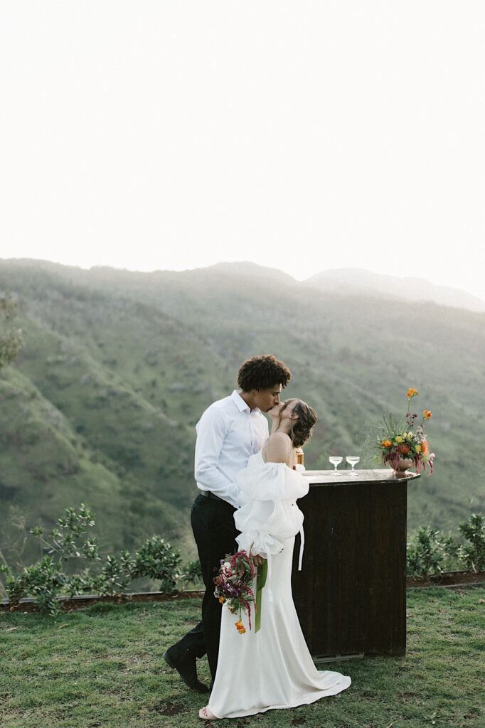A bride and groom kiss on their wedding day next to a mini bar with mountains behind them.