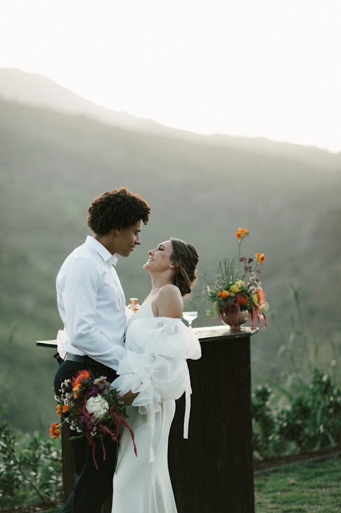 A bride and groom embrace on their wedding day next to a mini bar with mountains behind them.