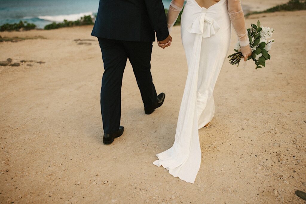 Waist down photo of a bride and groom walking away from the camera on Makapuu Beach after their elopement ceremony.