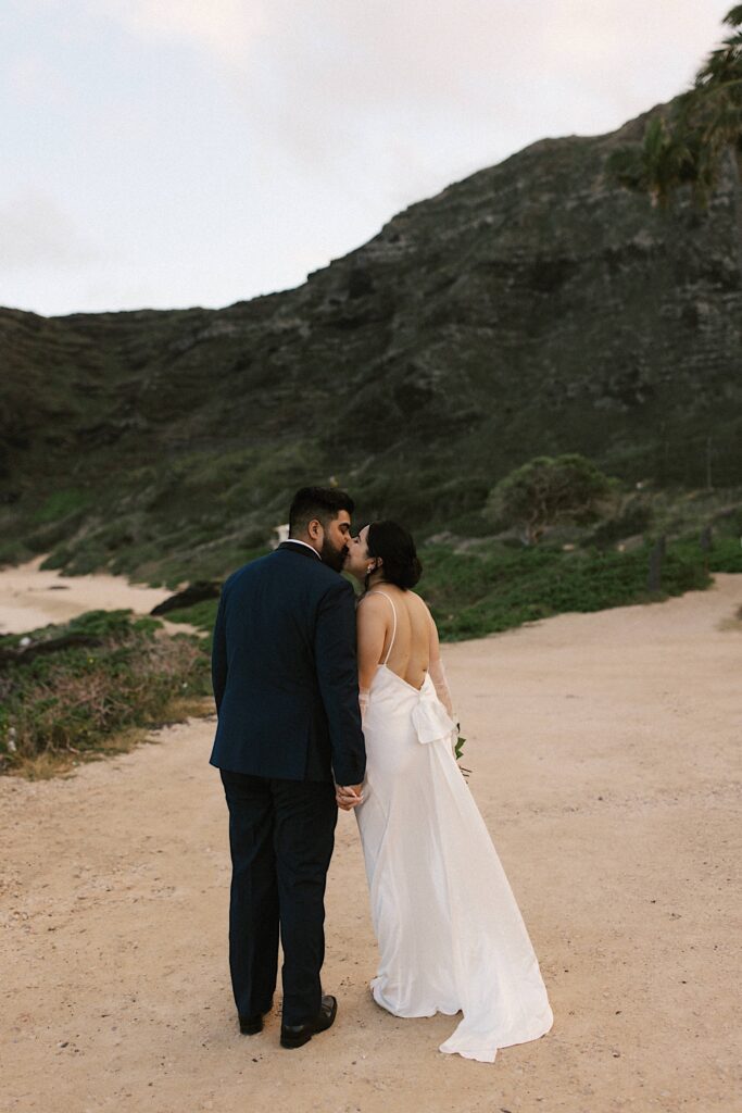 A bride and groom kiss on Makapuu Beach in Hawaii with their backs turned to the camera