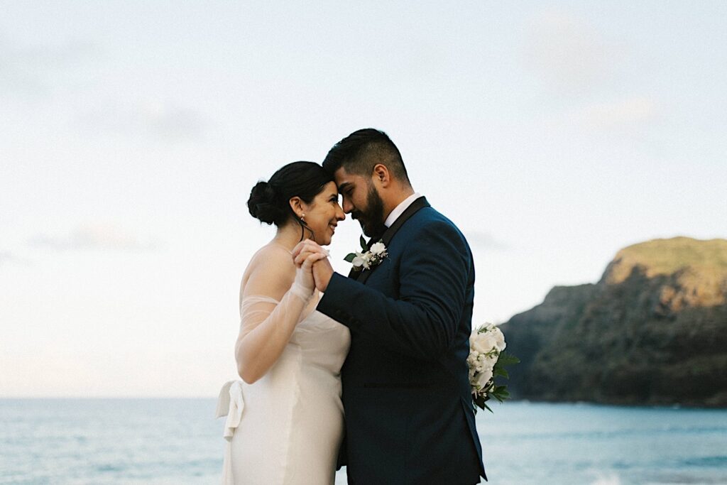 A bride and groom embrace one another and touch their foreheads together while on Makapuu Beach in Hawaii after their elopement ceremony