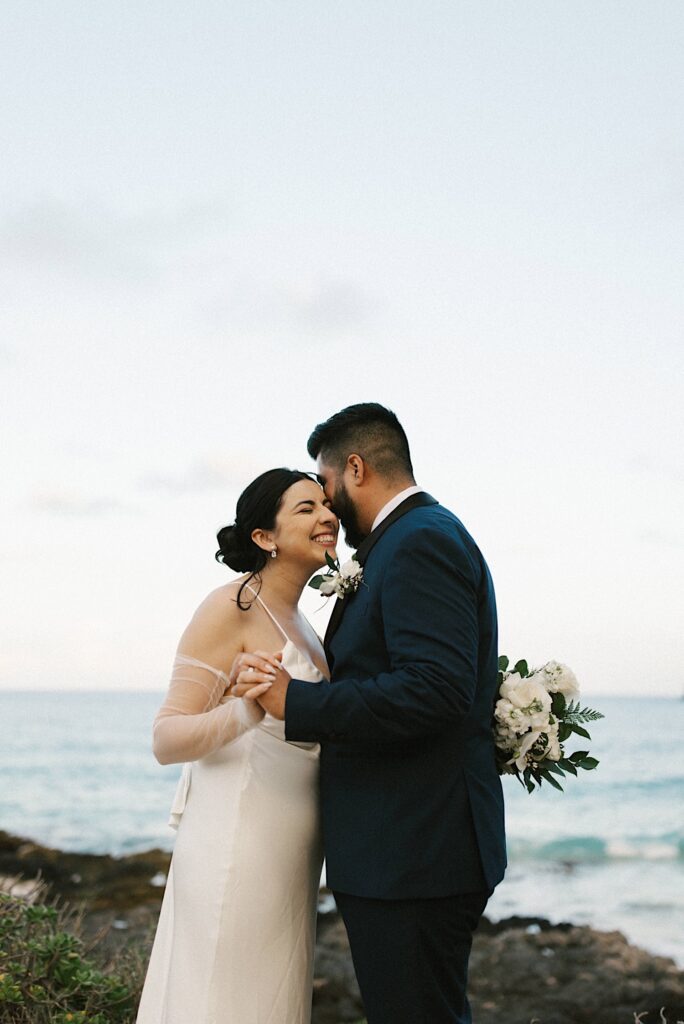 A bride and groom embrace one another on Makapuu Beach in Hawaii with the ocean behind them.