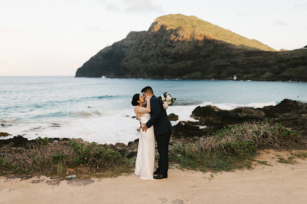 A bride and groom kiss while on Makapuu Beach in Hawaii after their elopement ceremony. The ocean and a mountain are behind them.