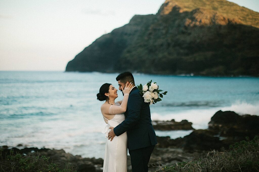 A bride and groom embrace while on Makapuu Beach in Hawaii after their elopement ceremony. The ocean and a mountain are behind them.