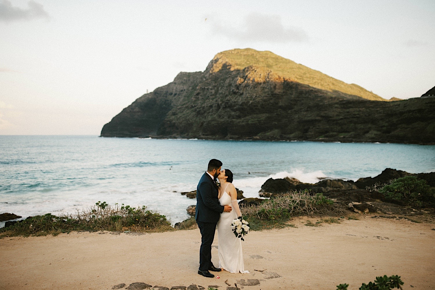 A bride and groom kiss while on Makapuu Beach in Hawaii after their elopement ceremony. The ocean and a mountain are behind them.