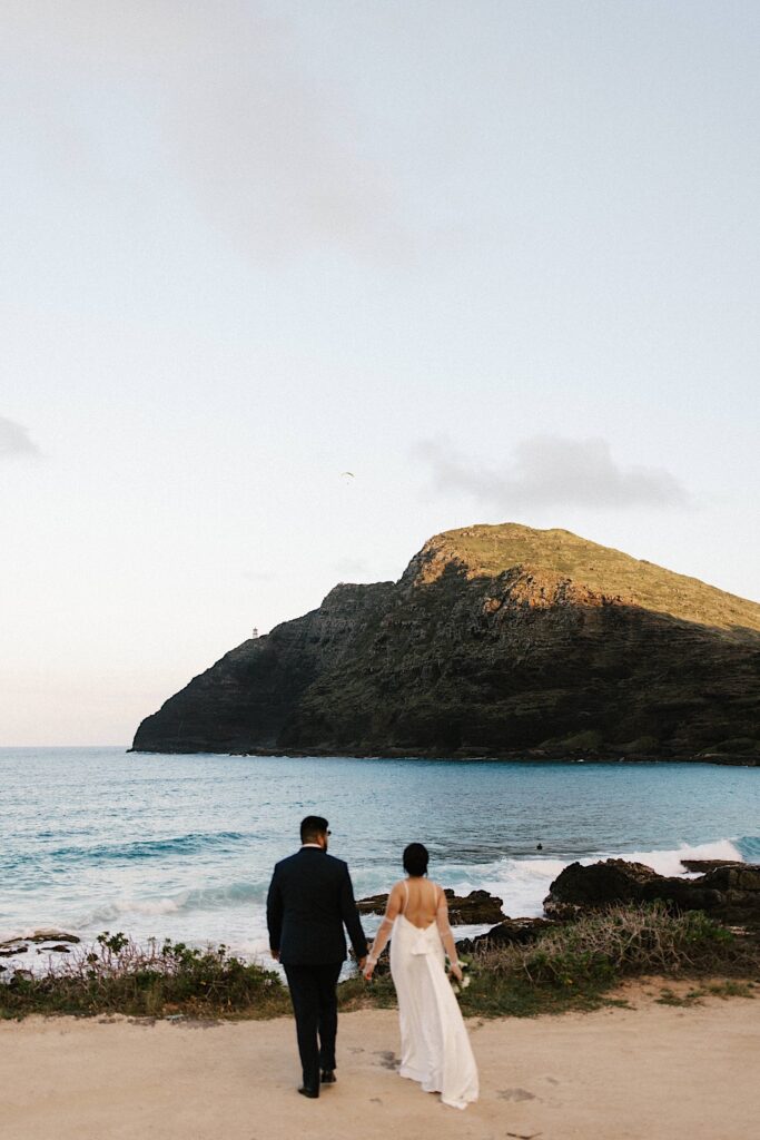 A bride and groom hold hands while looking out over the ocean and a mountain while on Makapuu Beach in Hawaii