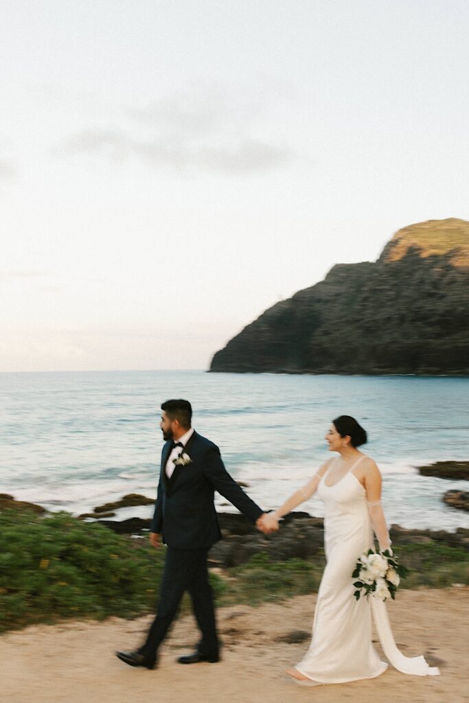 A bride and groom walk away from the ocean on Makapuu Beach while out of focus