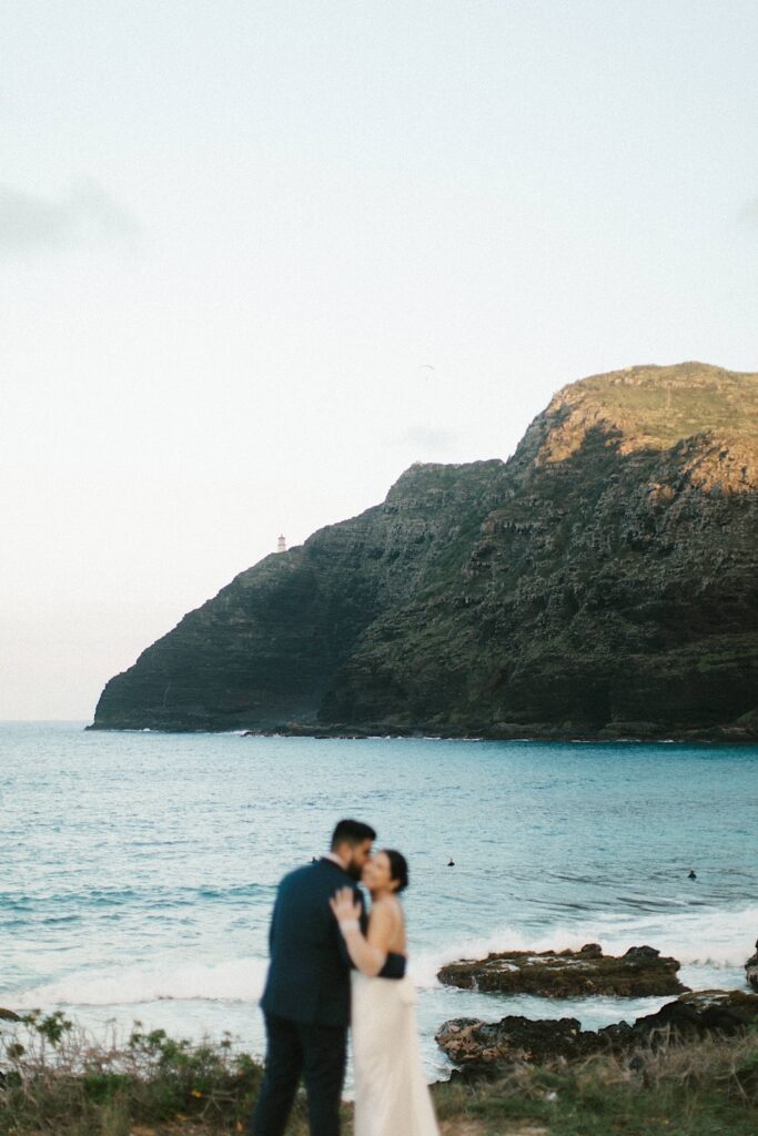 A bride and groom embrace one another on Makapuu Beach in Hawaii with the ocean and a mountain behind them.
