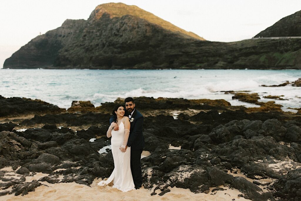 A bride and groom embrace one another and look at the camera on Makapuu Beach in Hawaii with the ocean and a mountain behind them after their elopement