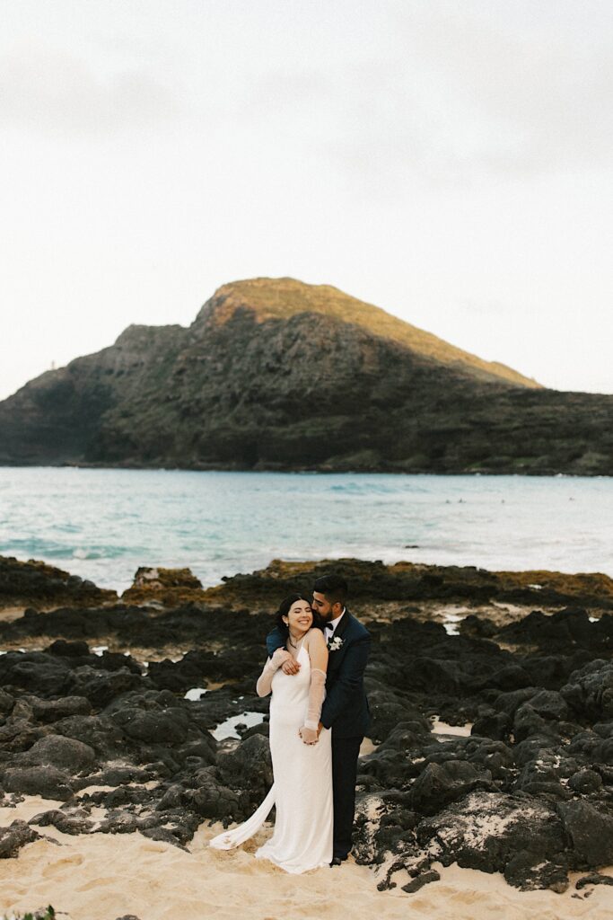 A bride and groom embrace one another on Makapuu Beach in Hawaii with the ocean and a mountain behind them.