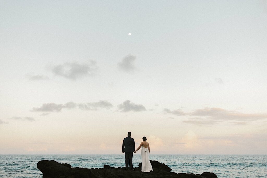 A bride and groom stand on Makapuu beach and hold hands looking out over the ocean with the moon above them after their elopement.