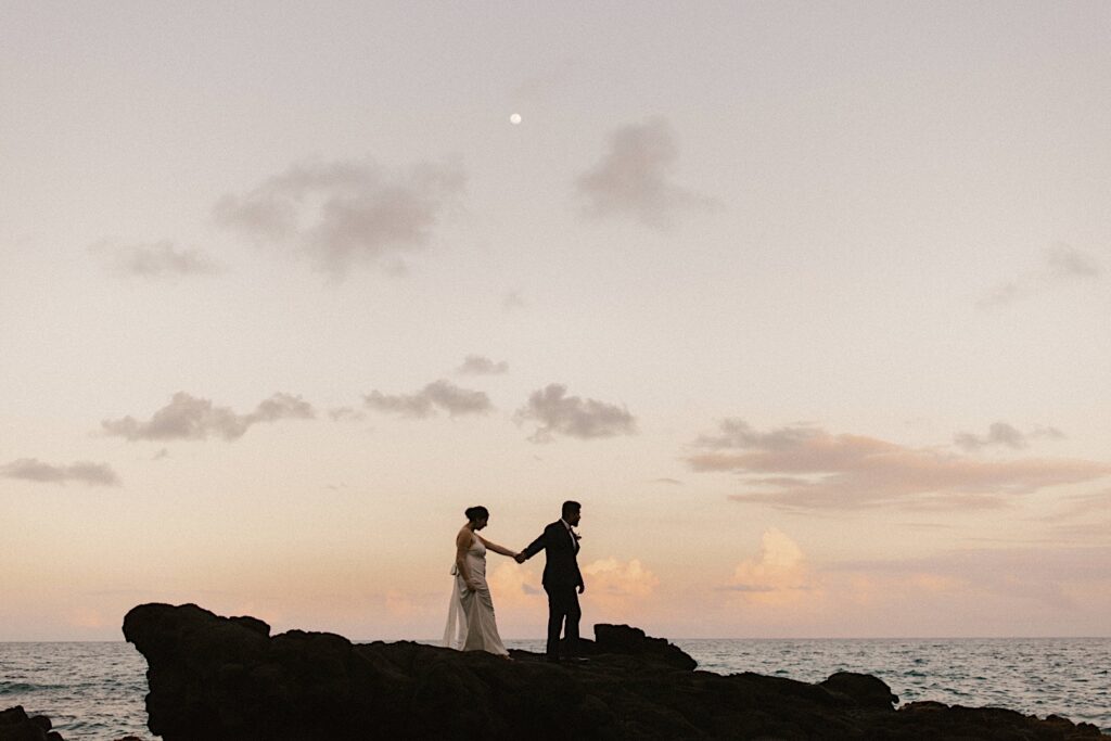 A bride and groom walk hand in hand on Makapuu beach with the ocean and sunset sky behind them on their elopement day.