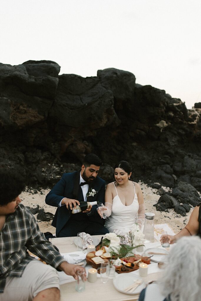 A bride and groom sit on Makapuu Beach and have a picnic together with their family after their elopement celebration.