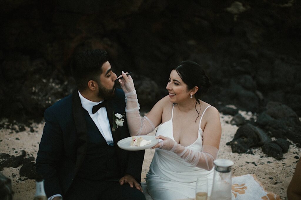 A bride feeds her groom a piece of cake while at their picnic on Makapuu Beach during their elopement celebration.