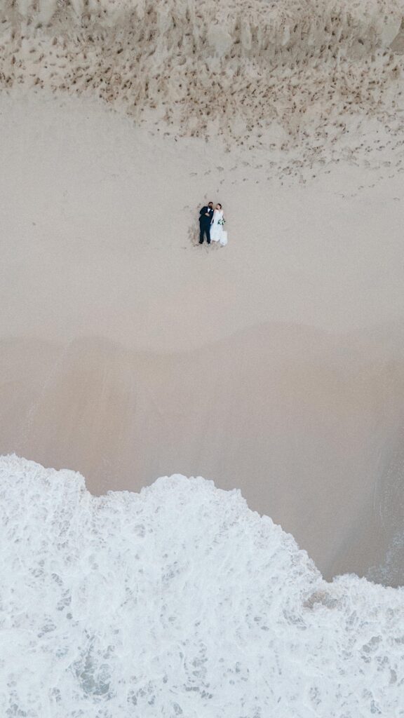 Drone shot of a bride and groom laying down in the sand together on Makapuu Beach while the water splashes near them.