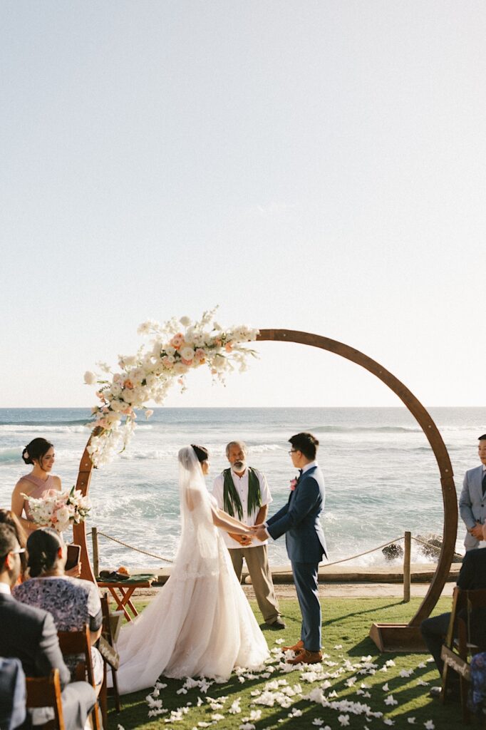 A bride and groom hold hands underneath their wedding arch which overlooks the ocean at Beach House in Kauai during their wedding ceremony