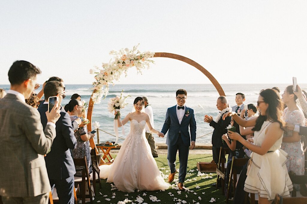 A bride and groom walk down the aisle after their wedding ceremony at their wedding venue Beach House in Kauai. The ocean and their wedding arch are in the background and guests are cheering them on.