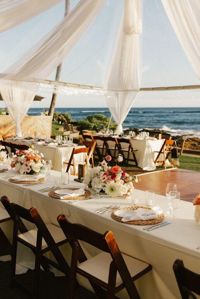 Tables underneath a tent are set up for a wedding reception with the ocean in the background.