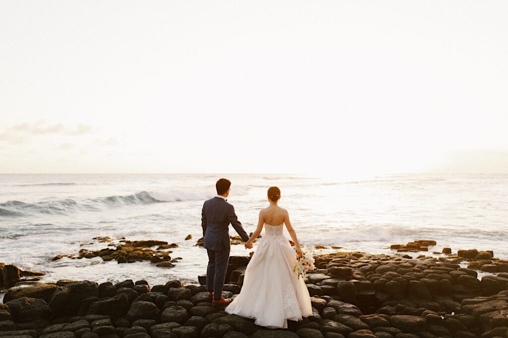 A bride and groom holding hands on the beach facing away from the camera, they're looking out at the ocean sunset at their wedding venue Beach House in Kauai