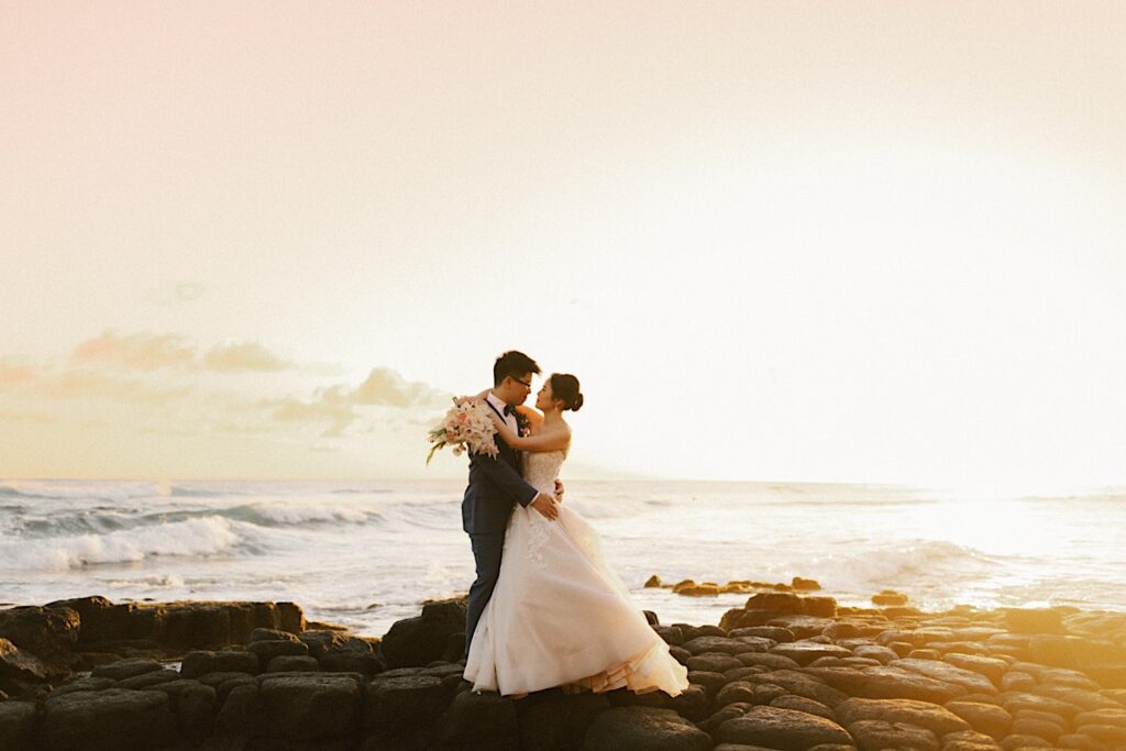 A bride and groom embrace on the beach looking at one another with the sunset and ocean behind them at their wedding venue Beach House in Kauai