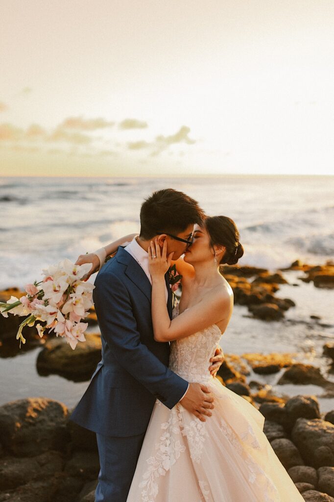 A bride and groom kiss on the beach with the ocean and a Hawaiian sunset in the background