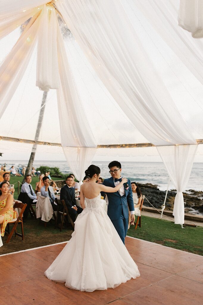 A bride and groom share their first dance underneath a tent with their wedding guests watching and the ocean in the background.