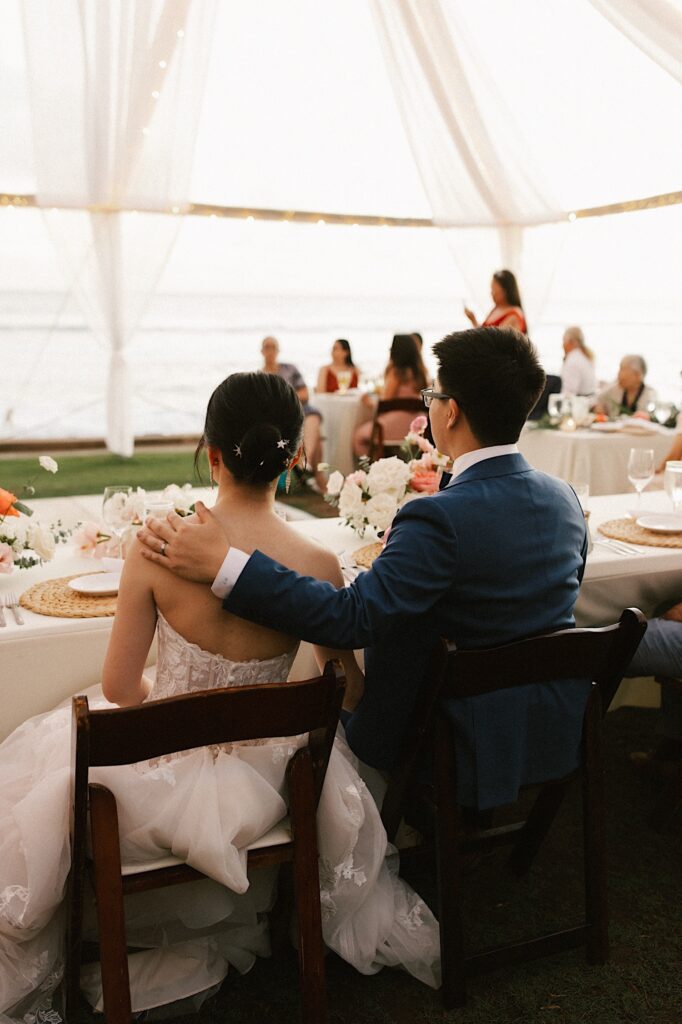 A bride and groom sit at a table during their wedding reception with their backs turned to the camera.
