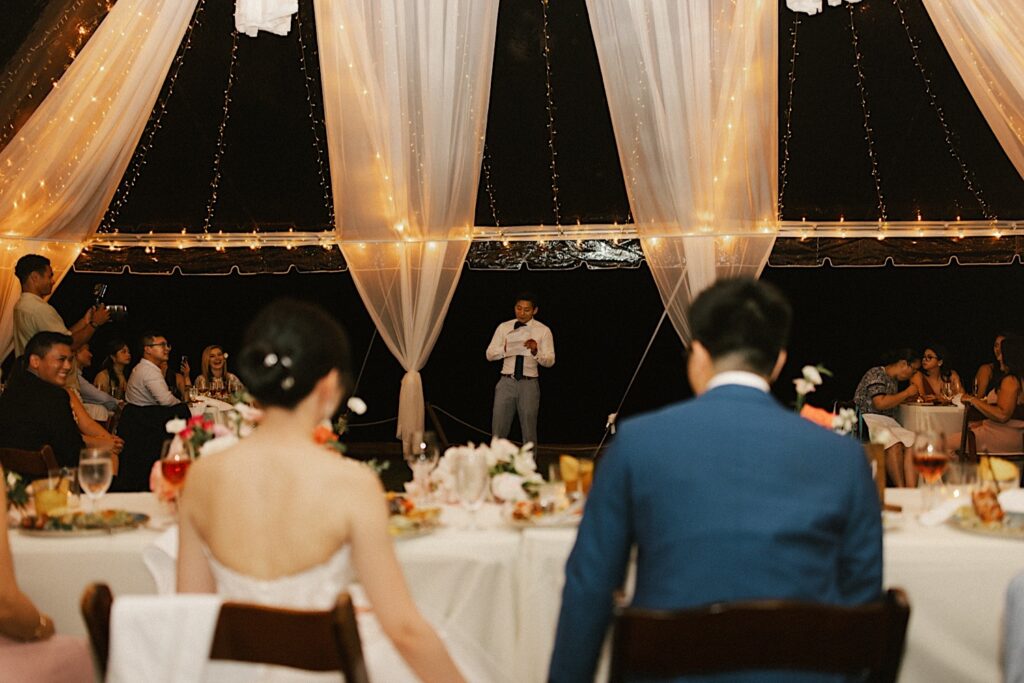 A man gives a speech at a wedding reception while the bride and groom sit in the foreground of the photo. The wedding reception is underneath at tent at the wedding venue Beach House in Kauai