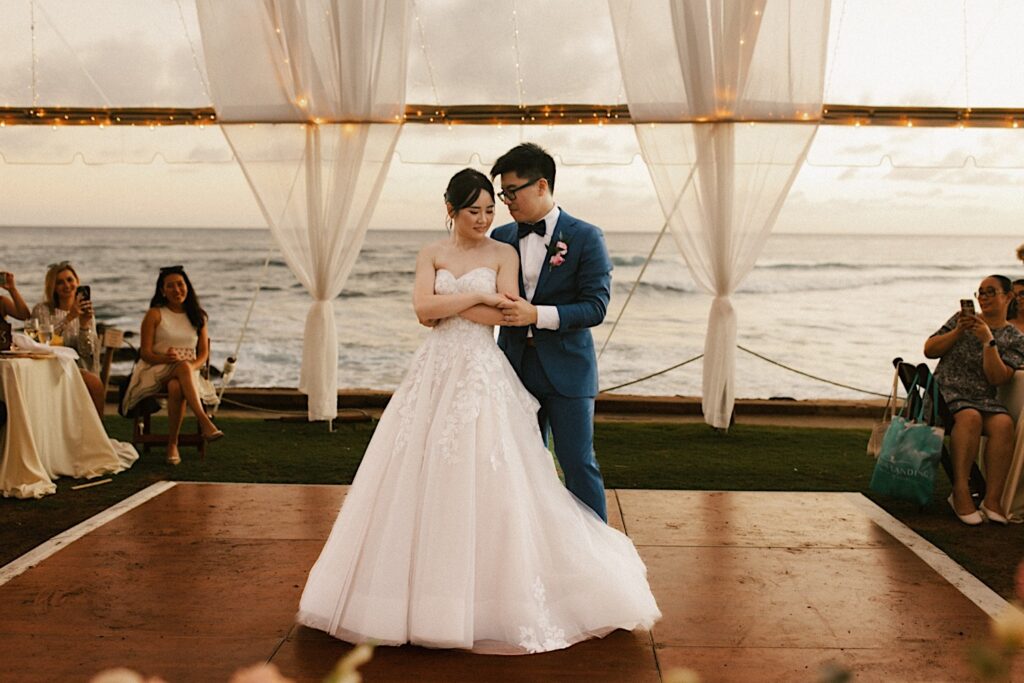 A bride and groom share their first dance at their wedding venue Beach House in Kauai. Their reception is underneath the tent at the venue with the ocean waves in the background.