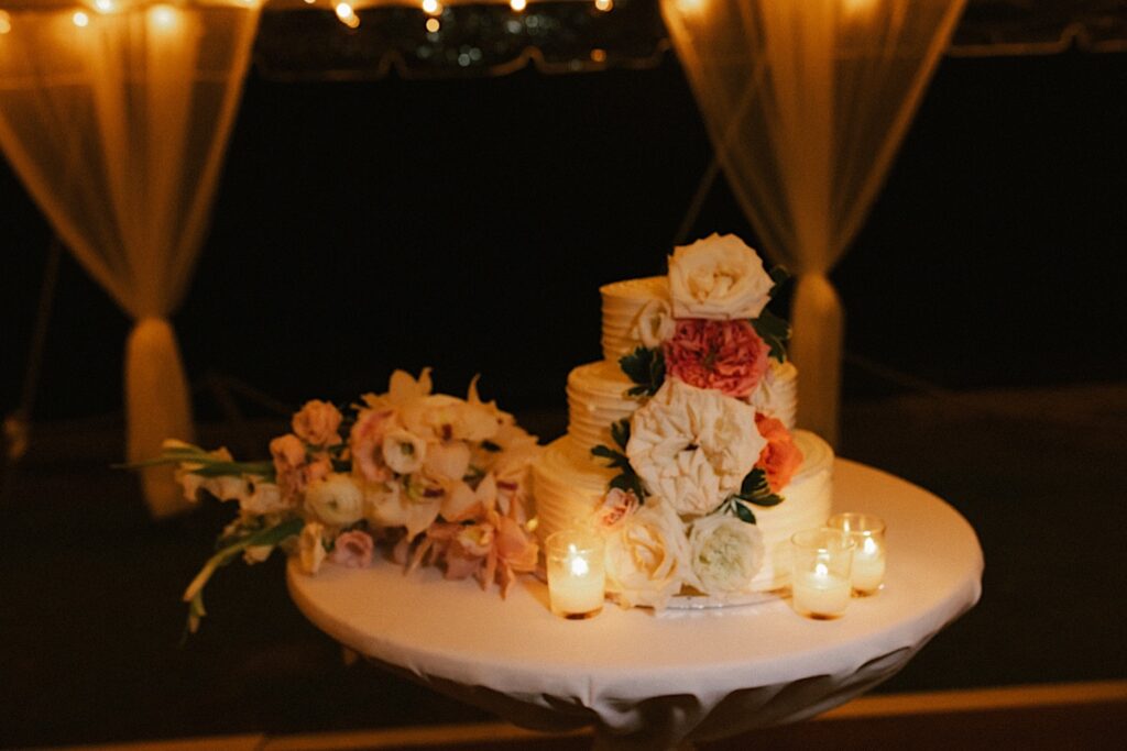 A wedding cake set up on a small white table with 3 candles and some florals next to it.