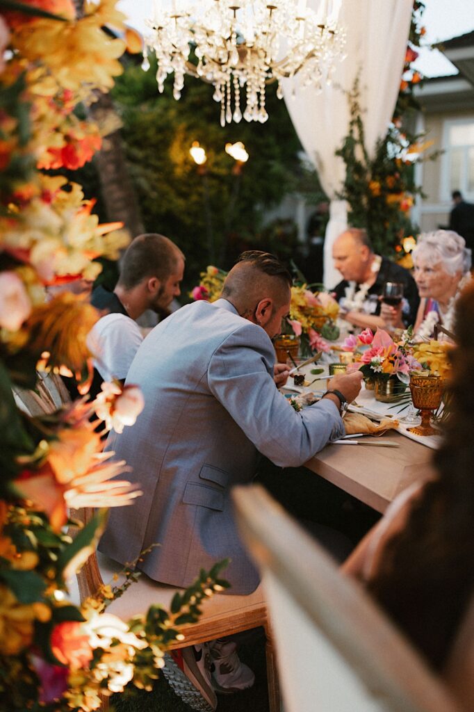 A groom and guests sit and eat at a table with florals and a chandelier above them.