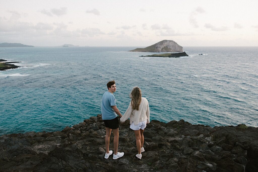 A couple stand on Makapuu Lookout in Hawaii looking out over the ocean and the islands, the man looks back at the camera nervous for his proposal
