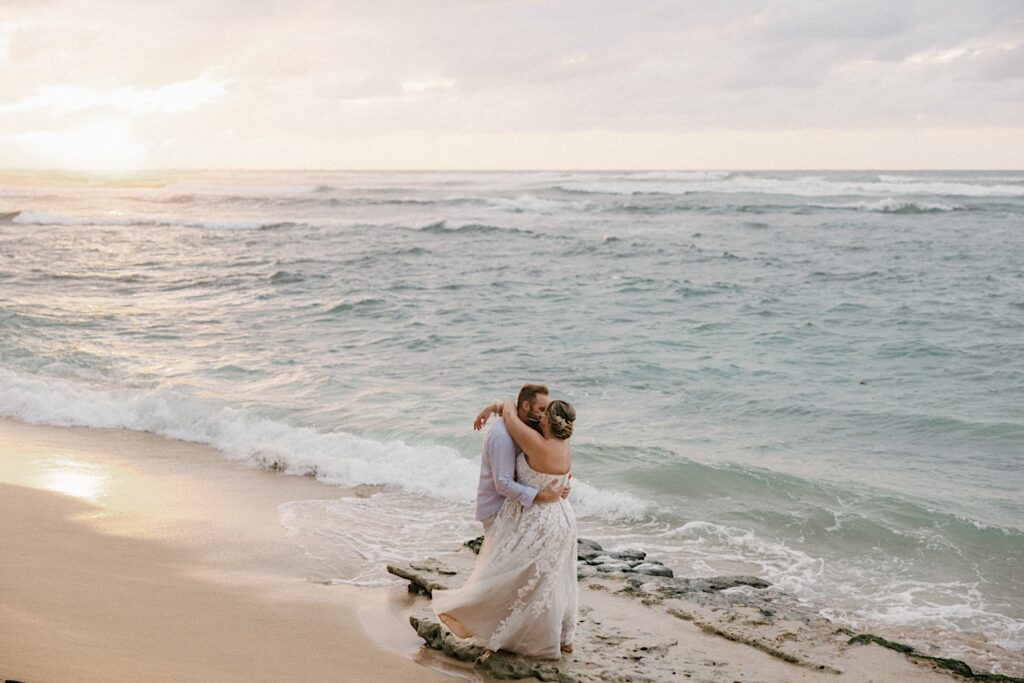 A couple kiss on the beach of Hawaii during their elopement, the sun is setting and waves are crashing behind them