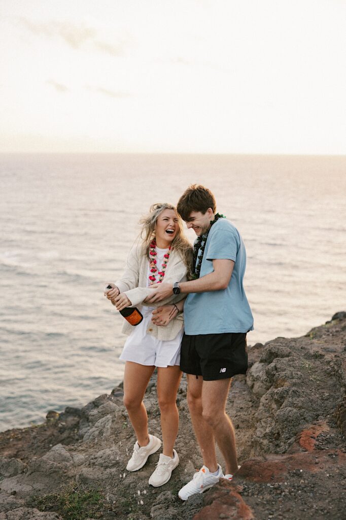 A couple laugh and smile on a cliff as they pop a bottle of champagne to celebrate their engagement