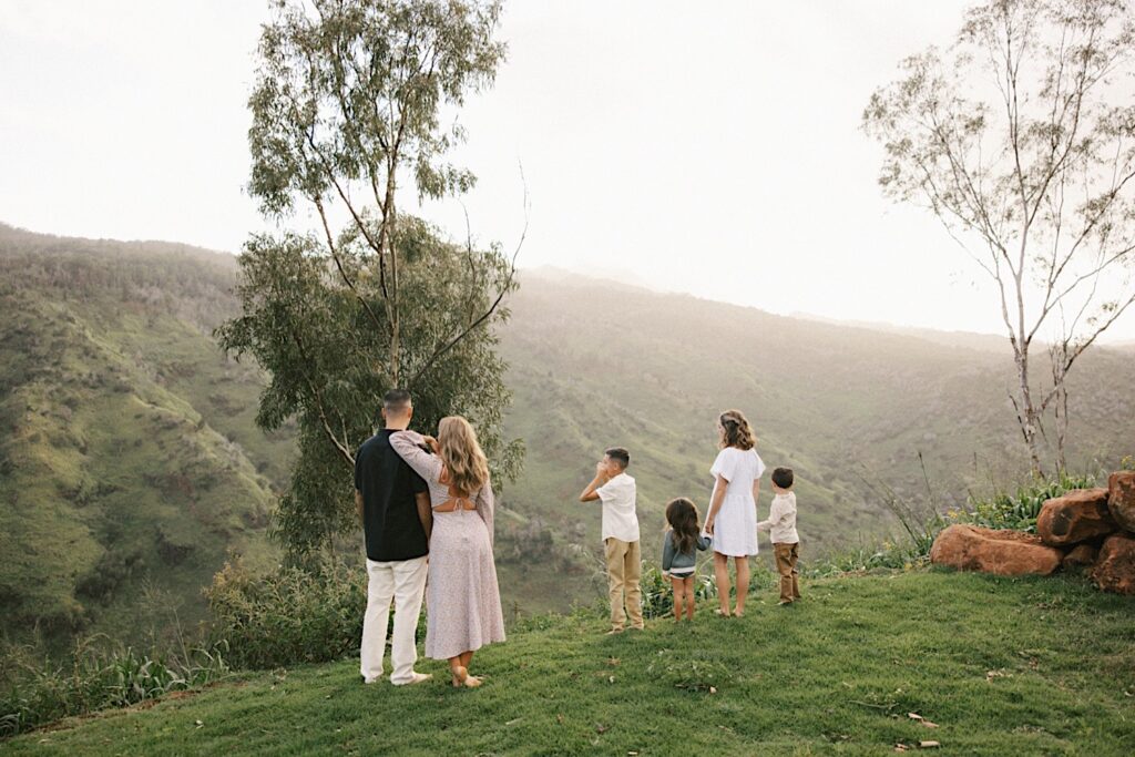 A man and woman stand side by side while their four children stand to the right of them holding hands, they all are looking out over a mountain during their family session on Oahu