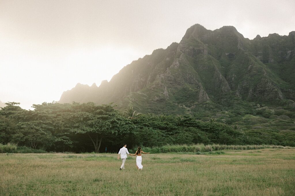 A couple walk hand in hand in a field towards a mountain in Hawaii during their engagement session