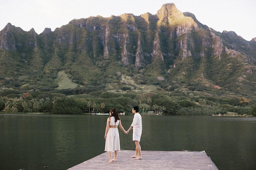 A couple in all white hold hands and stand on a dock looking out over the water and the mountains in the distance during their engagement session in Hawaii
